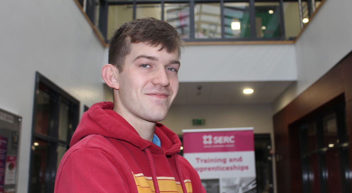 Male wearing red hoodie, smiling to camera pictured with apprenticeship poster in the background in college foyer